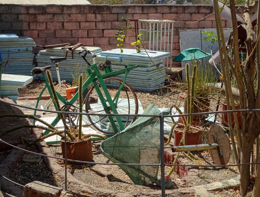 photograph of part of the Technology Demonstration Park at Vigyan Ashram, India. a green bicycle is visible, which has been converted into a tool that looks like a lathe. other tools and plants surround the bicycle and in the background there are materials such as stacks of styrofoam.