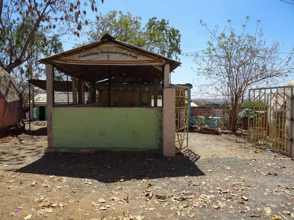photograph of the Technology Demonstration Park at Vigyan Ashram, India. there is a basic concrete building in the photo with a sign saying Technology Demonstration Park. the building and gates are painted green, pink and yellow. the sun is strong and casting shadows. the technologies displayed are visible in the background, as are other various buildings on the ashram.