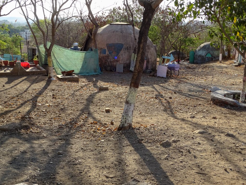 photograph of the yard area of Vigyan Ashram, India. the sun is lowering and creating strong shadows from the trees. a pink dome for shelter/housing is visible in the centre and another dome with a green triangle shaped door is visible further away. implements for plants such as plant pots and crates can be seen