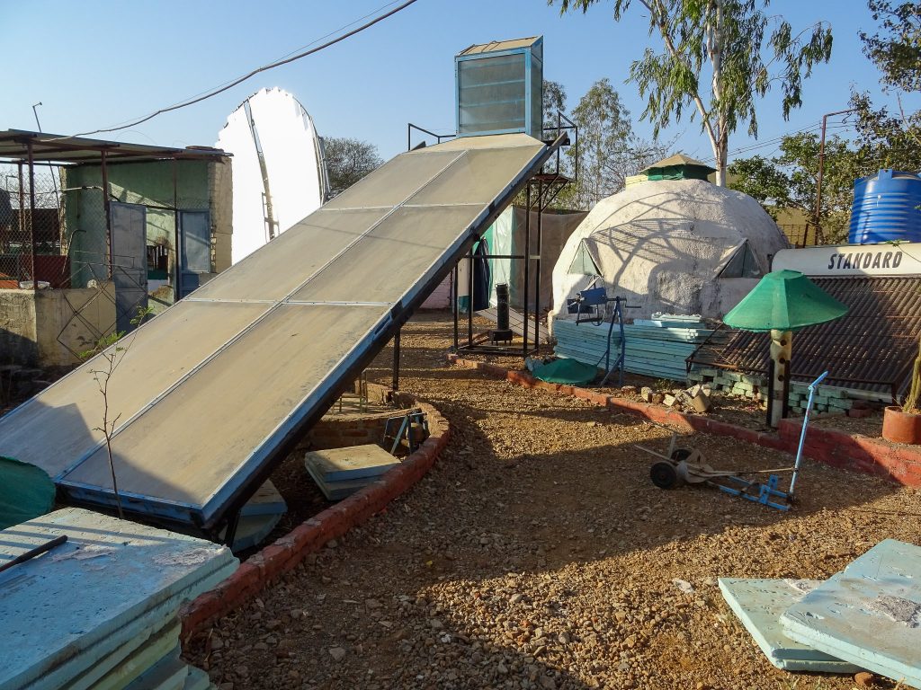 photograph of various inventions and prototypes displayed at the Technology Demonstration Park at Vigyan Ashram, India. visible is a large slanted vegetable dryer table, a large solar reflector disk, a dome for mushroom growing and various material stacks such as styrofoam and bricks.