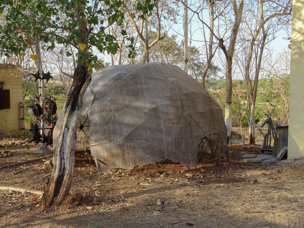 photograph of a geodesic dome being built in Vigyan Ashram, India. the dome structure is in place and is covered with a slightly transparent textile. there are trees behind the dome and a couple of buildings visible on either side.