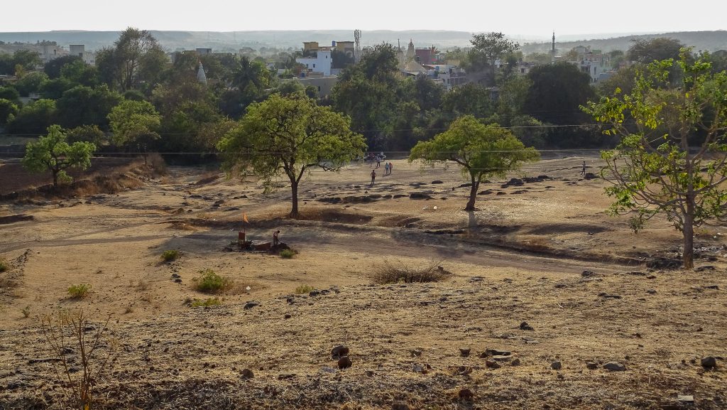 a landscape looking from Vigyan Ashram towards the village of Pabal in India. the earth is dry and brown and ochre with grasses. at the end of a slop some figures are visible, playing football. a row of trees are leafy and green and within and beyond the trees some buildings are visible, mosque towards, a communications tower. the scene extends further into a misty landscape of soft hills. the atmosphere is calm and quiet and rural.