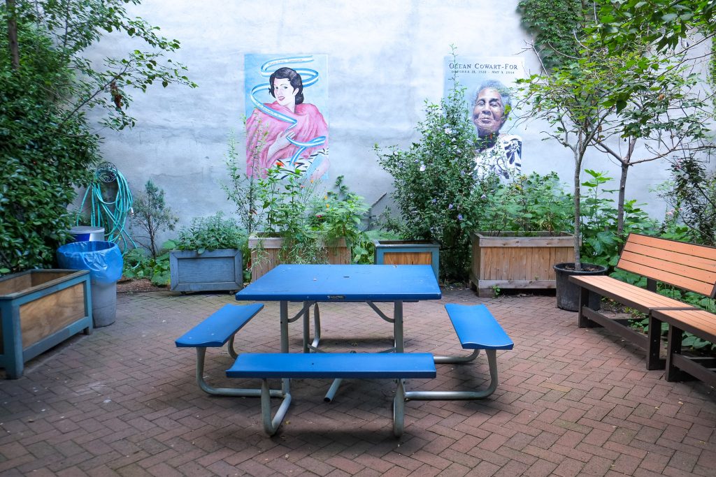 photograph of part of a community garden in New York's Lower East Side. there are two frescos or murals of people painted on a building wall. plant boxes line the wall and a bright blue picnic table with attached benches is in the middle. wooden benches mark the right side of the sitting space, which is tiled with stone flooring.