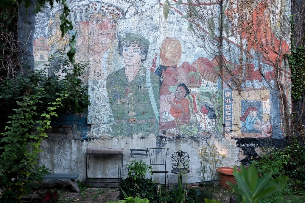 photograph of a wall in a community garden in Manhattan's Lower East Side, New York City. the wall has a large mural painted on it, in soft tones of green and ochre, depicting different types of people, from soldiers to African women holding a child. three metal garden chairs are in a very graceful row along the wall. the tonal varieties are all subdued and in harmony, creating a relaxed atmosphere.