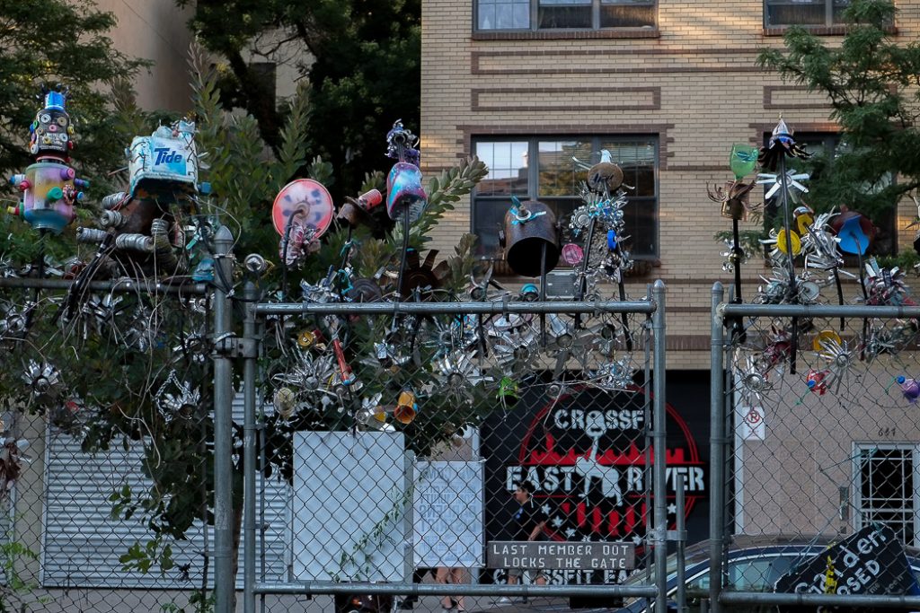 photograph of decorations along a fence of a community garden in Manhattan's Lower East Side, New York City. the decorations are made mainly of metal and stick up and down off the top of the fence looking like a bunch of whirligigs. there are other creations too, made of waste such as detergent boxes and a robot made of waste materials.