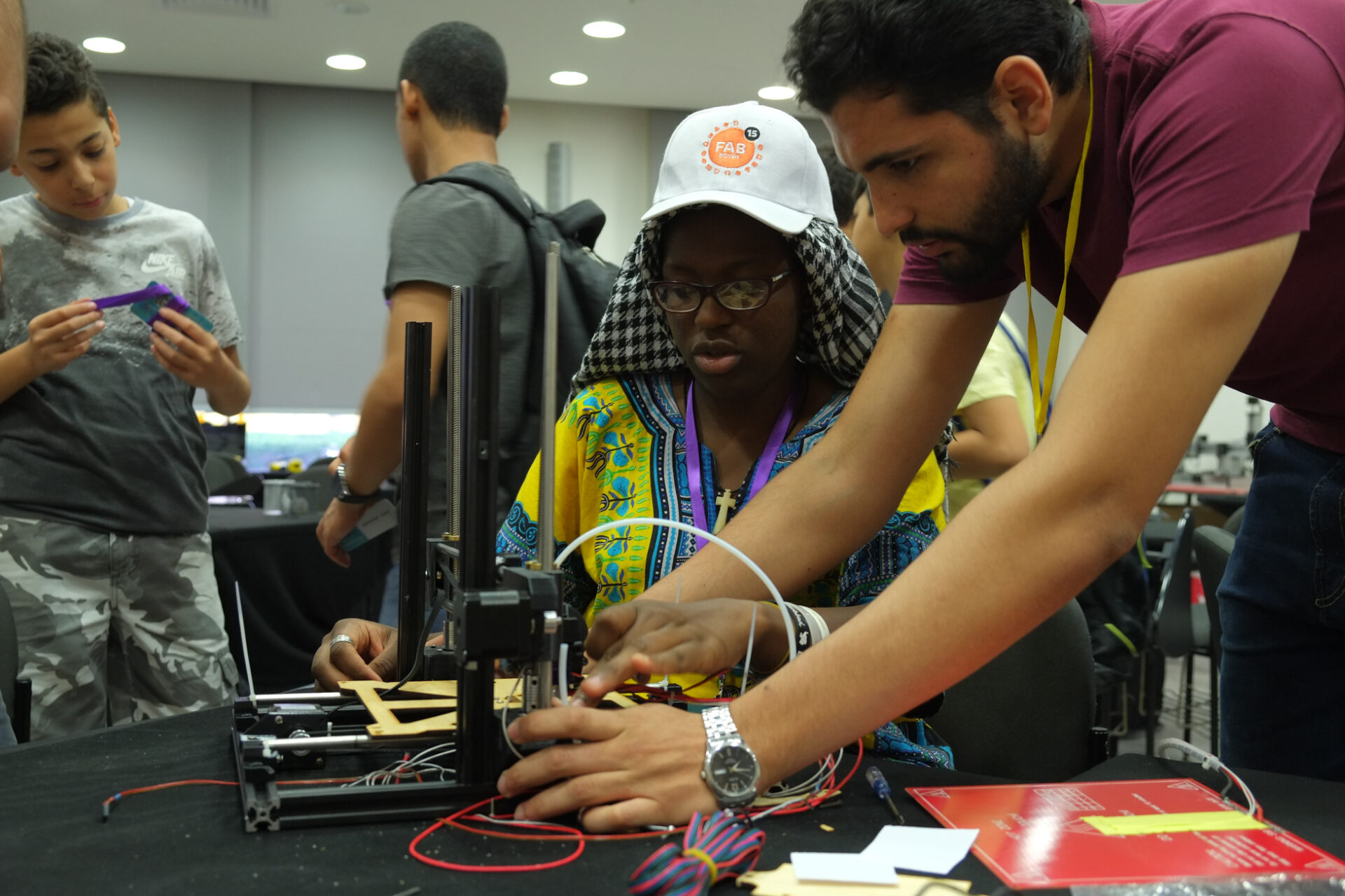 photograph of a group of people-of-colour makers building 3D printers. In the foreground a young man and a young woman wearing a FAB15 baseball cap are looking at the printer they are building together and have their hands on parts putting it together. There are wires, metal and plastic parts visible. In the background two other people are visible looking at the work going on in the room or at printed parts.
